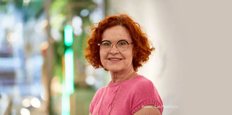 Een vrouw met rood krullend haar en een bril, gekleed in een roze trui, die glimlacht in een moderne omgeving.