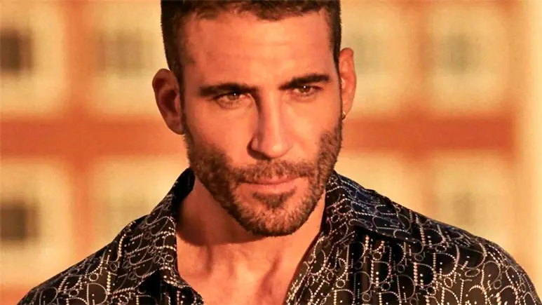 Close-up van Miguel Angel Silvestre met baard en een shirt met print, tegen een onscherpe stedelijke achtergrond.