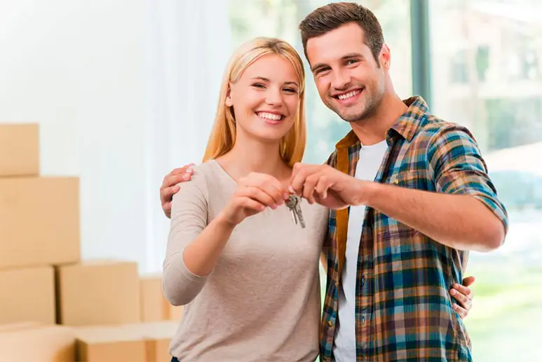 Pareja sonriente sosteniendo las llaves de su nuevo apartamento frente al mar, con cajas de mudanza en el fondo.