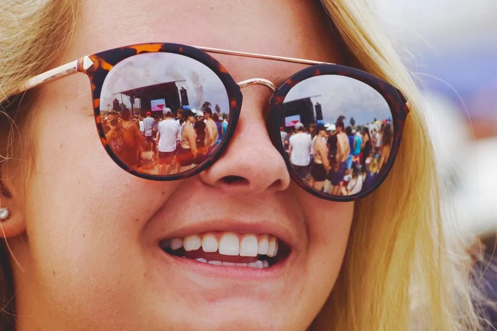 Primer plano de una chica sonriendo en un festival, con el escenario y el público reflejados en sus gafas de sol