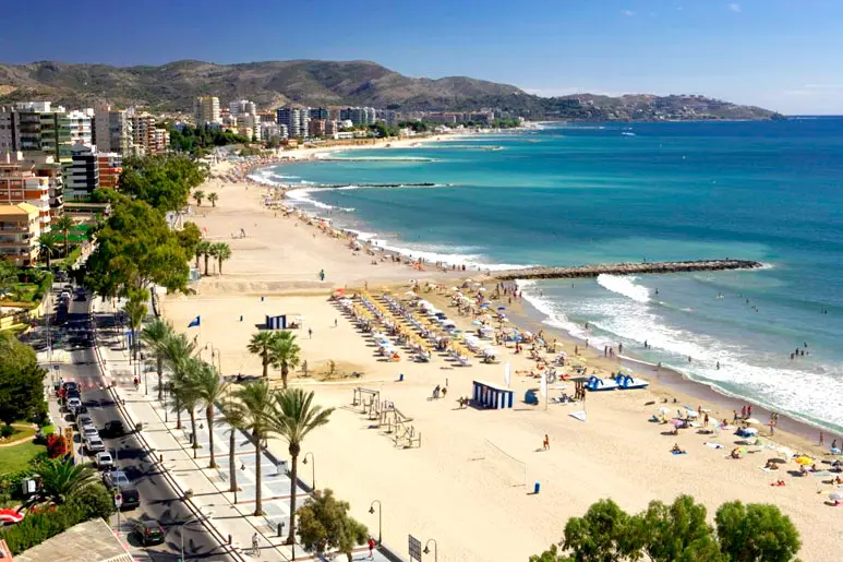 Panoramablick auf den Strand Heliópolis in Benicàssim mit goldenem Sand, Sonnenschirmen, der Strandpromenade und dem Mittelmeer unter einem klaren Himmel.